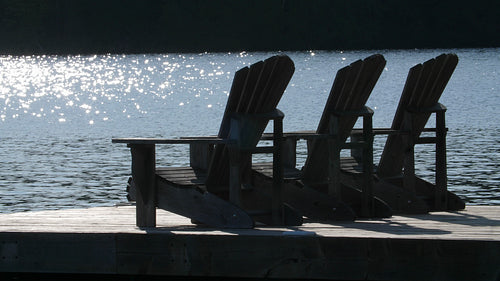 Three Muskoka chairs in silhouette. Sparkling lake. Cottage country. 4K.