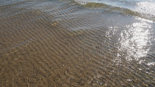 Slow motion sunlit lake water and wave with rippled sand pattern below. HD video.