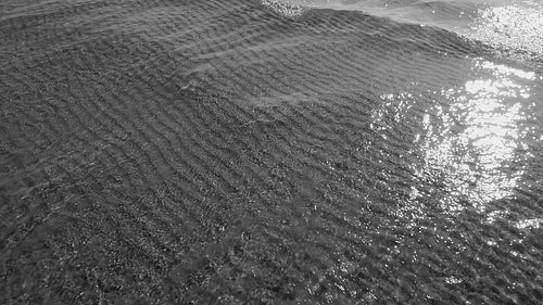 Slow motion sunlit lake water and wave with rippled sand pattern below. Black and white. HD video.