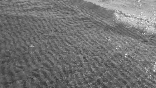 Tracking slow motion sunlit lake wave in shallow water. Rippled sand. Black and white. HD stock video.
